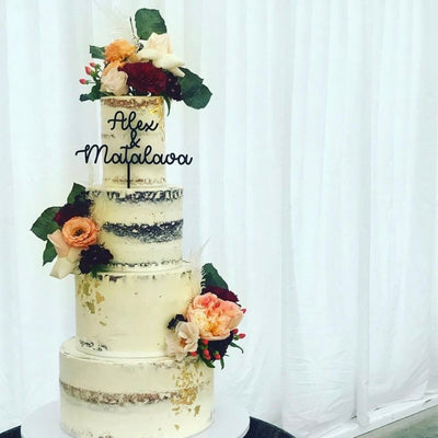 A four tier white wedding cake, accompanied with florals, and a black acrylic cake topper saying Alex and Matalowa.