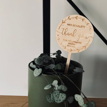 A green potted plant with heart-shaped leaves sits on a wooden surface beside a Personalised Wreath Teacher Planter engraved, "Mrs Scarlett thank you for helping me grow, Indie 2020." A black railing is visible in the background.