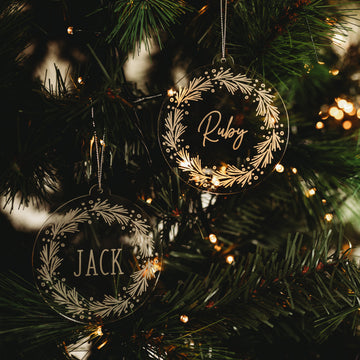 Two Pine Frosted Christmas Baubles, each personalized with JACK or Ruby, hang on a tree with warm white string lights illuminating the green pine branches. The clear acrylic ornaments feature charming white wreath designs.