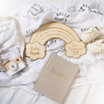 A flat lay featuring a Personalised Rainbow Announcement Plaque with "Baby Mann" and "Due November 2019," a beige bump book, eco-friendly bamboo toy camera, baby shoes, and Hello World blanket.