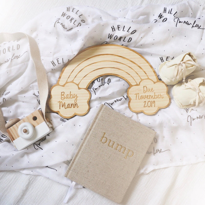 A flat lay featuring a Personalised Rainbow Announcement Plaque with "Baby Mann" and "Due November 2019," a beige bump book, eco-friendly bamboo toy camera, baby shoes, and Hello World blanket.