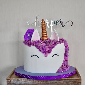 A unicorn-themed cake with purple flowers, a gold horn, ears, closed eyes, a purple number 4, and a Mirror Personalised Cake Topper reading Harper sits on a purple polka-dot ribbon cake board.