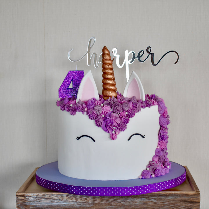 A unicorn-themed cake with purple flowers, a gold horn, ears, closed eyes, a purple number 4, and a Mirror Personalised Cake Topper reading Harper sits on a purple polka-dot ribbon cake board.