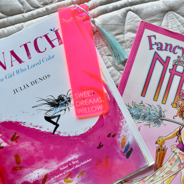 A neon pink see through acrylic bookmark with sleepy eyelashes etched on the end, a light blue tassel, and the words Sweet Dreams, Willow etched on the bottom. Sitting atop a children&