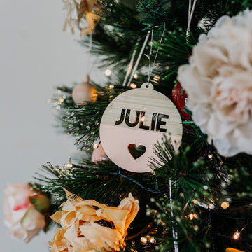The Original Personalised Christmas Bauble, featuring the name JULIE and a heart cutout, hangs on a decorated tree with lights and flowers, adding a special touch to your family’s Christmas tradition.