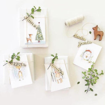 A collection of personalized swing tags with various designs, including names and festive illustrations like Santa and reindeer, displayed on a white surface with greenery accents.