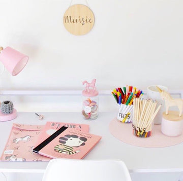 A tidy white desk with pink accents holds notebooks, a jar of candy, colorful pens, pencils, wooden animal figures, and a pink lamp. Above hangs the Personalised Natural Names Plaque—perfect personalised decor for any space.