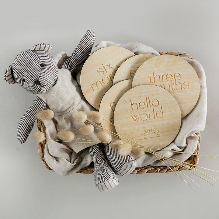 A woven basket with a bear, bunny tails and round bamboo milestone cards saying hello world, and the months of the year. Hello World has a sunshine engraved on it.