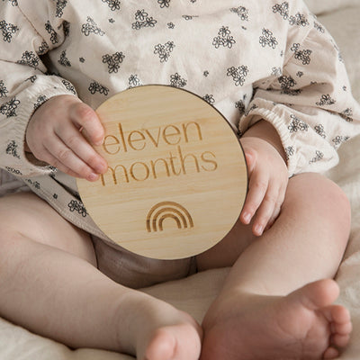 A baby holding a round bamboo milestone plaque saying eleven months, and engraved with a rainbow.