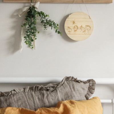 A round light bamboo plaque with a childs drawing of 4 people and a sun engraved on it. Hanging above a bed next to a flower arrangement. 