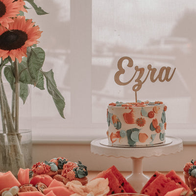 A light bamboo cake topper saying Ezra, sitting atop a white cake with orange, blue and green buttercream paint strokes. Sitting on a party table with florals and food.